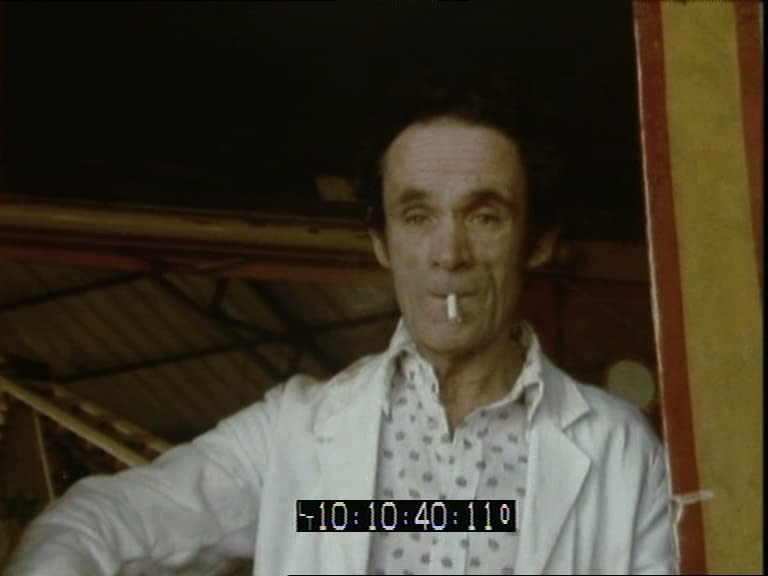 A video still showing a man in a white coat, with a white cigarette drooping from his mouth.